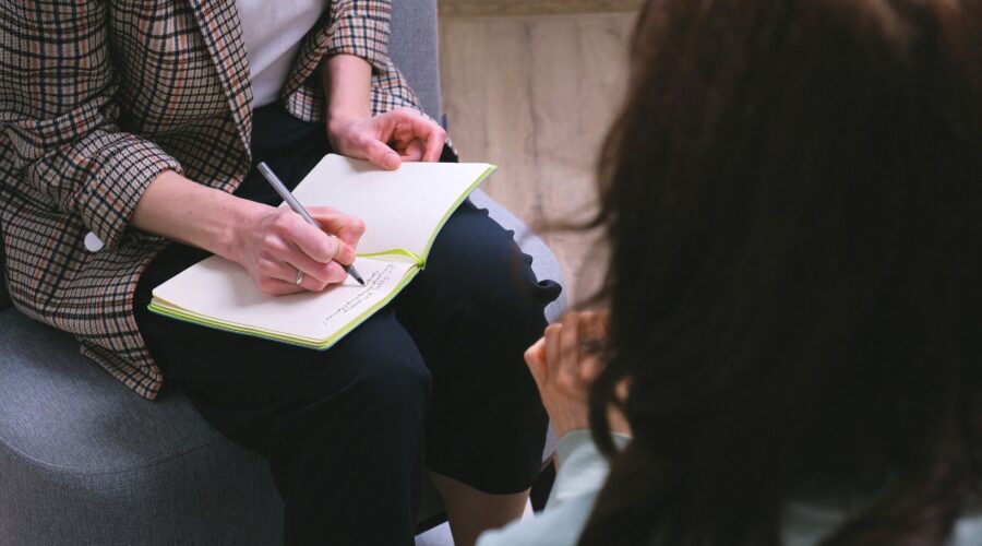 A therapist taking notes while sitting across from a client during a therapy session, highlighting personalized care and a goal-oriented approach to enhancing the therapy experience at Embodied Wellness Center.