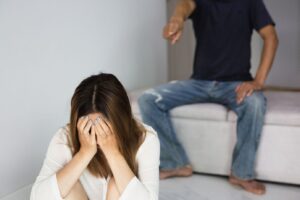 A distressed woman sits with her head in her hands while a man yells at her in the background, illustrating the impact of verbal abuse. This image represents emotional distress, manipulation, and the importance of setting boundaries in unhealthy relationships.