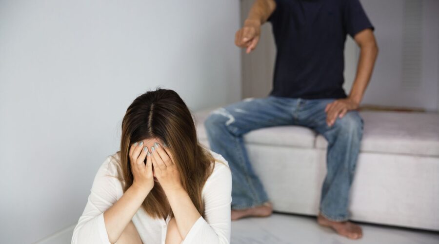 A distressed woman sits with her head in her hands while a man yells at her in the background, illustrating the impact of verbal abuse. This image represents emotional distress, manipulation, and the importance of setting boundaries in unhealthy relationships.